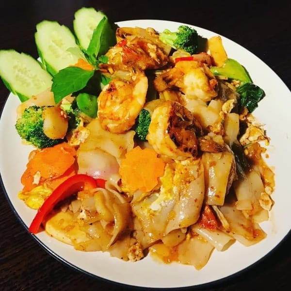 Thai drunken noodles with shrimp, broccoli, carrots, and cucumbers on a white plate.